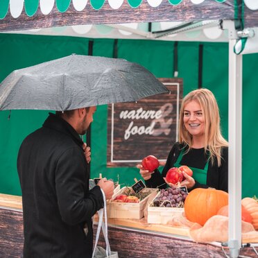 The saleswoman in the market tent holds various fruits in her hand. The customer is standing in front of the sales tent at the counter.